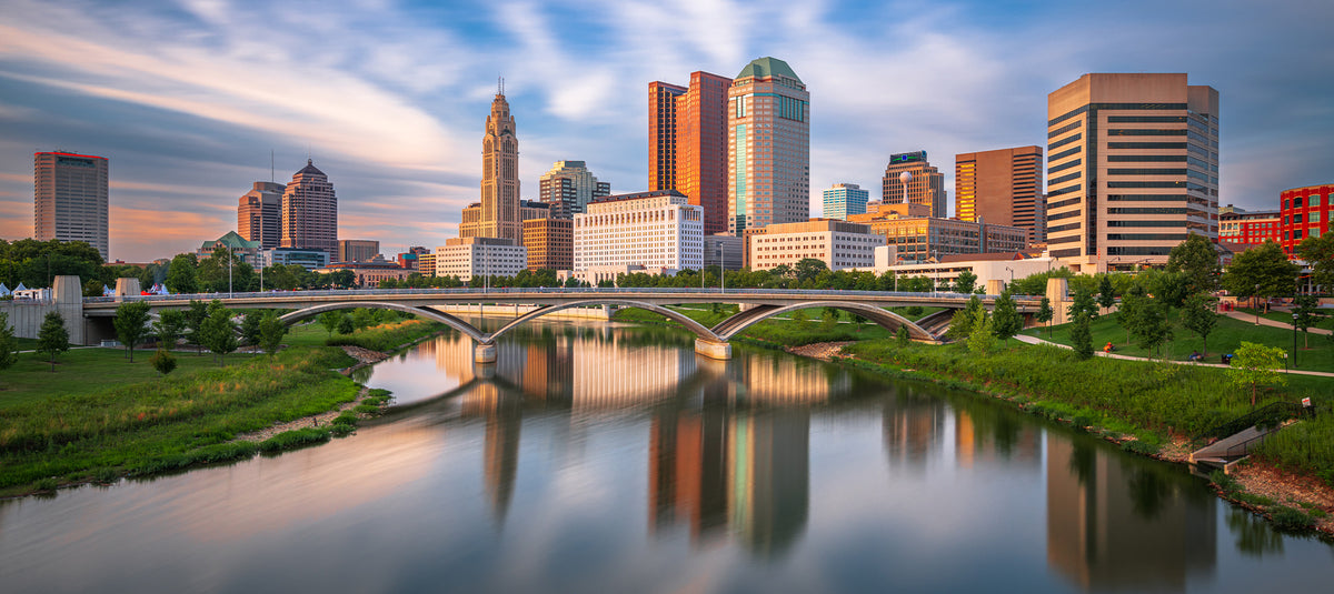 Panoramic view of the Columbus, Ohio skyline reflecting over the Scioto River with arched bridges and waterfront parklands