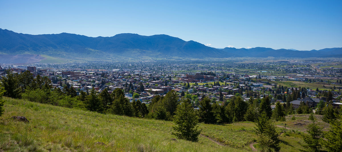 Panoramic landscape view of a Montana valley town with rolling green hills and distant mountain ranges under a clear blue sky